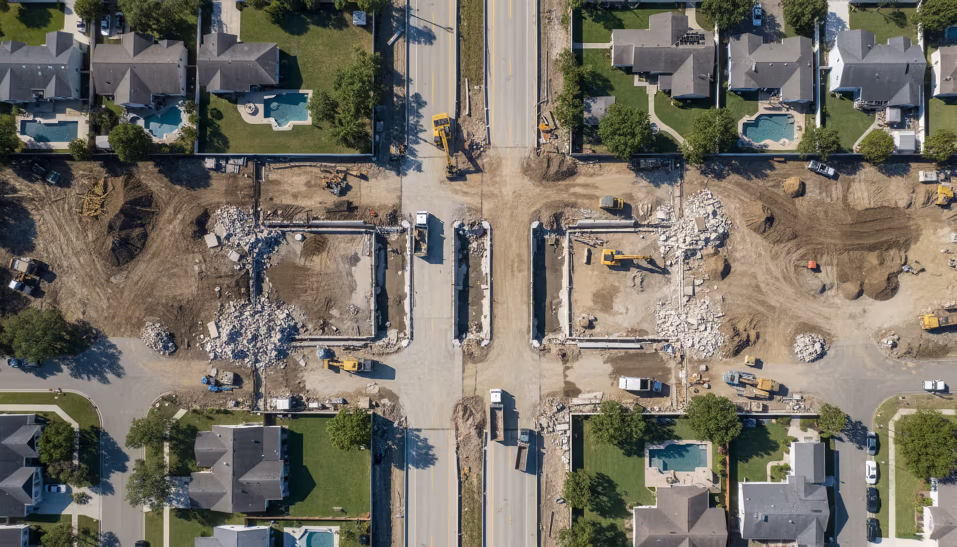 Aerial view of highway construction cutting through suburban neighborhood with partially demolished houses and heavy machinery